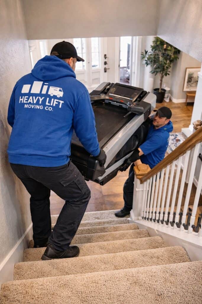 Two professional movers from Heavy Lift Moving Co safely carrying a treadmill downstairs inside a Utah home using proper lifting techniques.